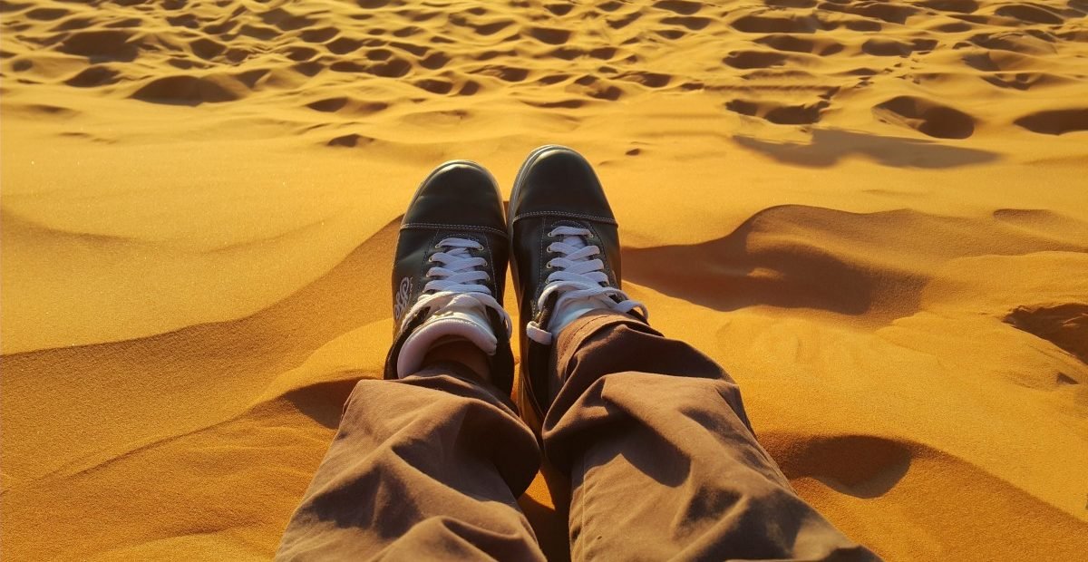 traveler sitting on golden Moroccan sand dunes at sunset near Safi and Essaouira, feet in black sneakers visible in foreground