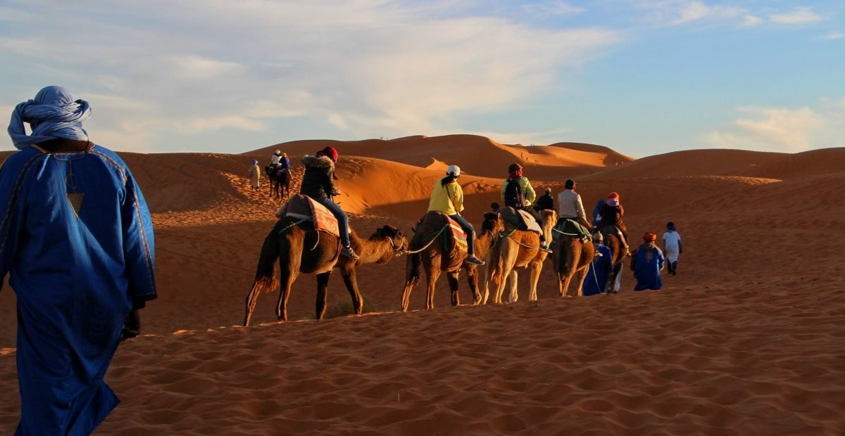 camel caravan with tourists guided by locals in traditional blue robes crossing the golden sand dunes of the Sahara Desert at sunset in Morocco