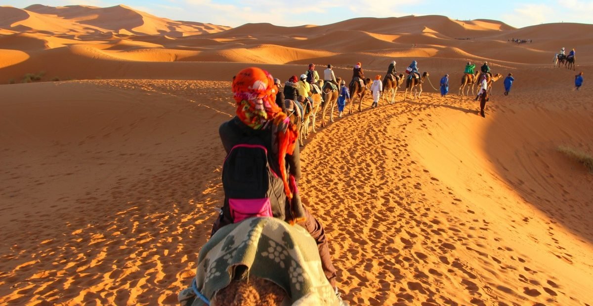 camel caravan with tourists riding through golden Sahara Desert sand dunes at sunset in Morocco, guided by locals in traditional blue attire