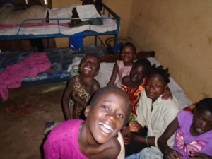 group of smiling Ugandan children sitting together in a modest dormitory room at Mercy Hearts Uganda orphanage school, with bunk beds and simple bedding