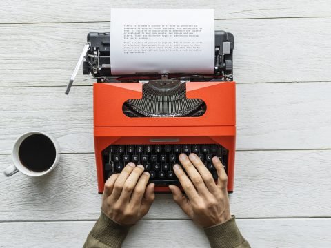 Hands typing on a red typewriter beside a cup of coffee on a white wood desk