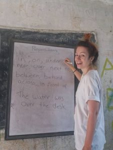 Volunteer teacher in Nepal smiles while giving an English lesson on prepositions at a rural classroom with a chalkboard.