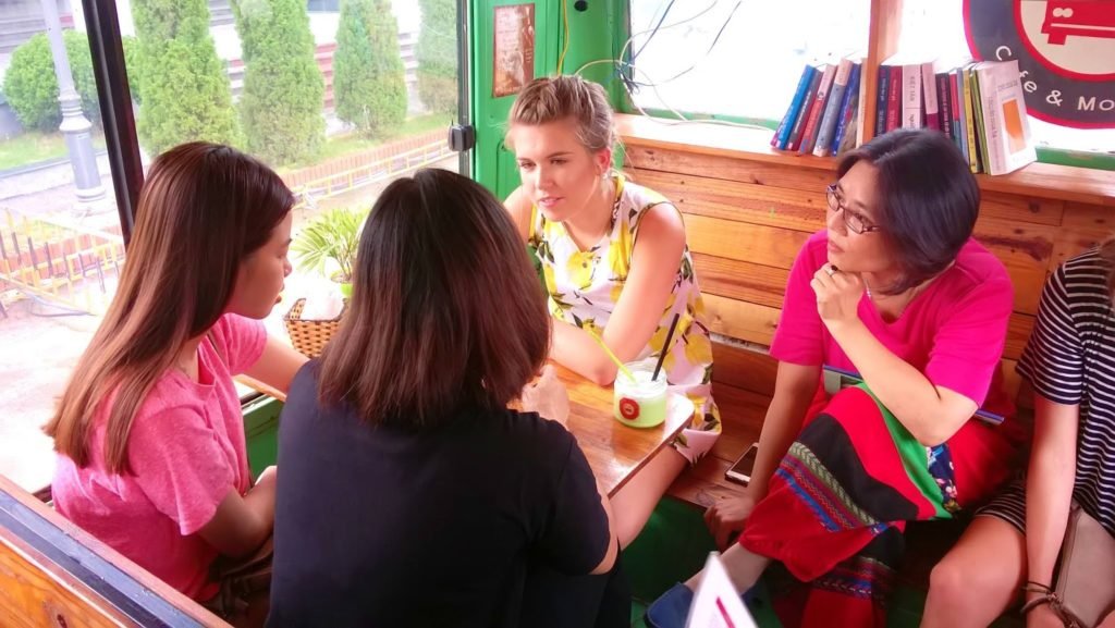 International volunteer teaching English in Hanoi café setting, engaged in conversation with local Vietnamese students as part of a cultural exchange program.
