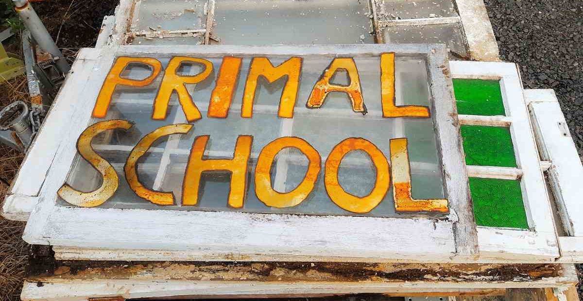 Old wooden window frame repurposed as a hand-painted sign with the words “Primal School” in bold yellow and orange letters.