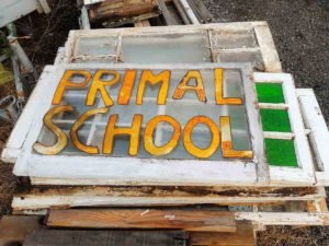 Old wooden window frame repurposed as a hand-painted sign with the words “Primal School” in bold yellow and orange letters.