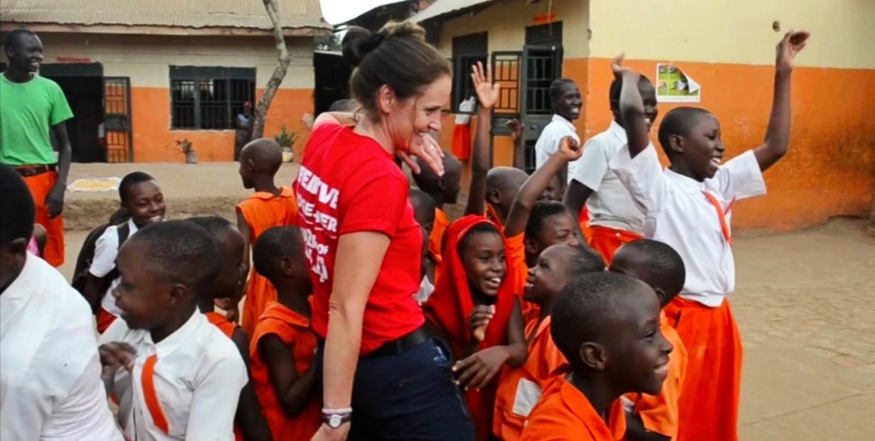 female volunteer playing and smiling with Ugandan school children in orange uniforms at an orphanage school courtyard in Kampala, Uganda