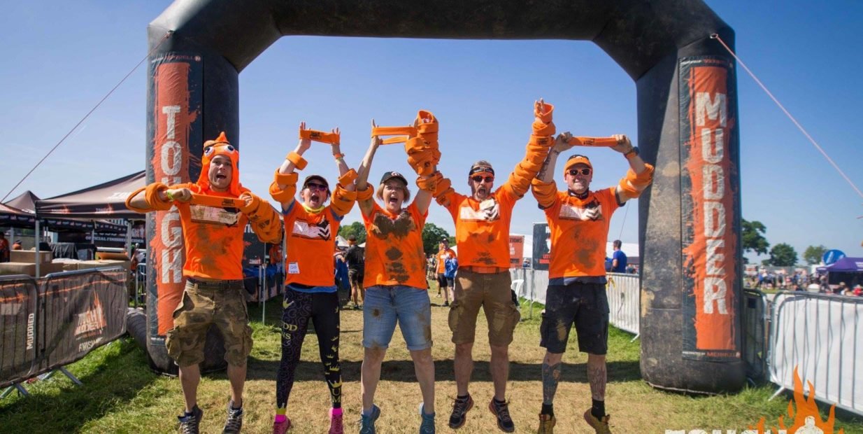 Five Tough Mudder volunteers in bright orange shirts jump and celebrate under a black Finish arch at a muddy outdoor course.