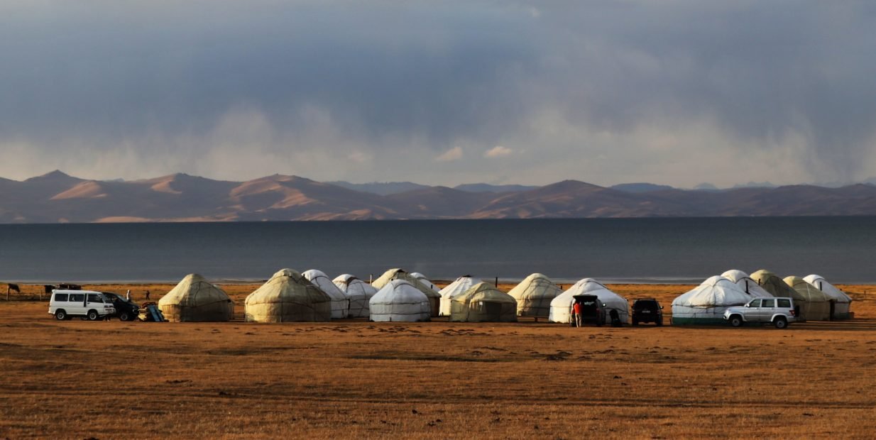 Series of yurts (traditional felt-covered tents) set up near a lake in the Kyrgyz steppe, with mountains in the background - scene typical of guesthouse or yurt camp networking in rural Kyrgyzstan.