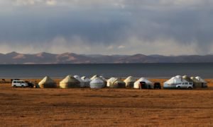 Series of yurts (traditional felt-covered tents) set up near a lake in the Kyrgyz steppe, with mountains in the background - scene typical of guesthouse or yurt camp networking in rural Kyrgyzstan.