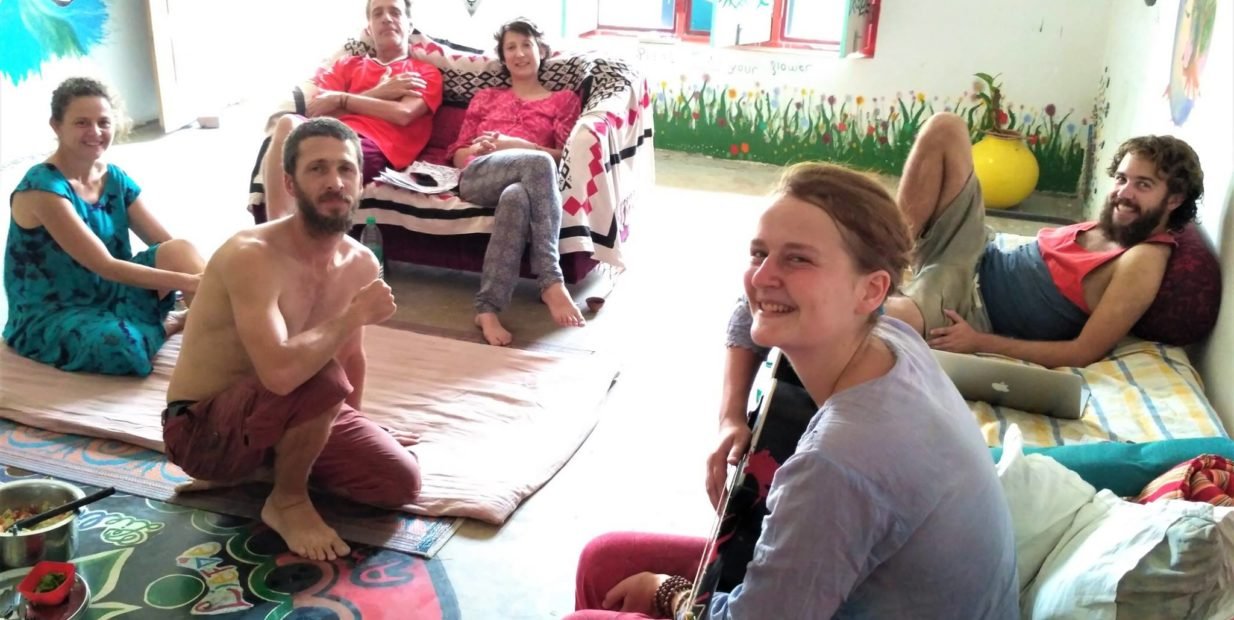A group of international volunteers relaxing in a colorful, artistic communal space at an eco-village in Varanasi, India. Some are sitting on mats and sofas, one person holds a guitar, surrounded by wall murals and natural light—showing the creative, peaceful community vibe.