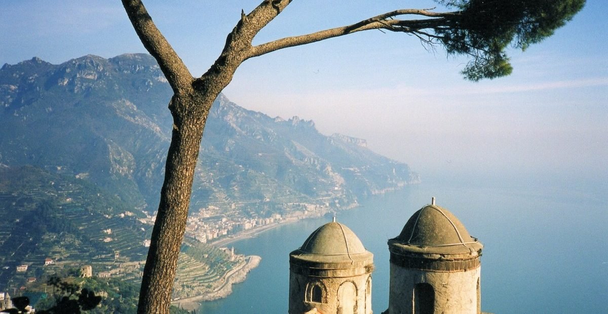 Panoramic view of Ravello, Italy from Villa Rufolo with Mediterranean sea, mountain cliffs, and historic domed towers under a pine tree.