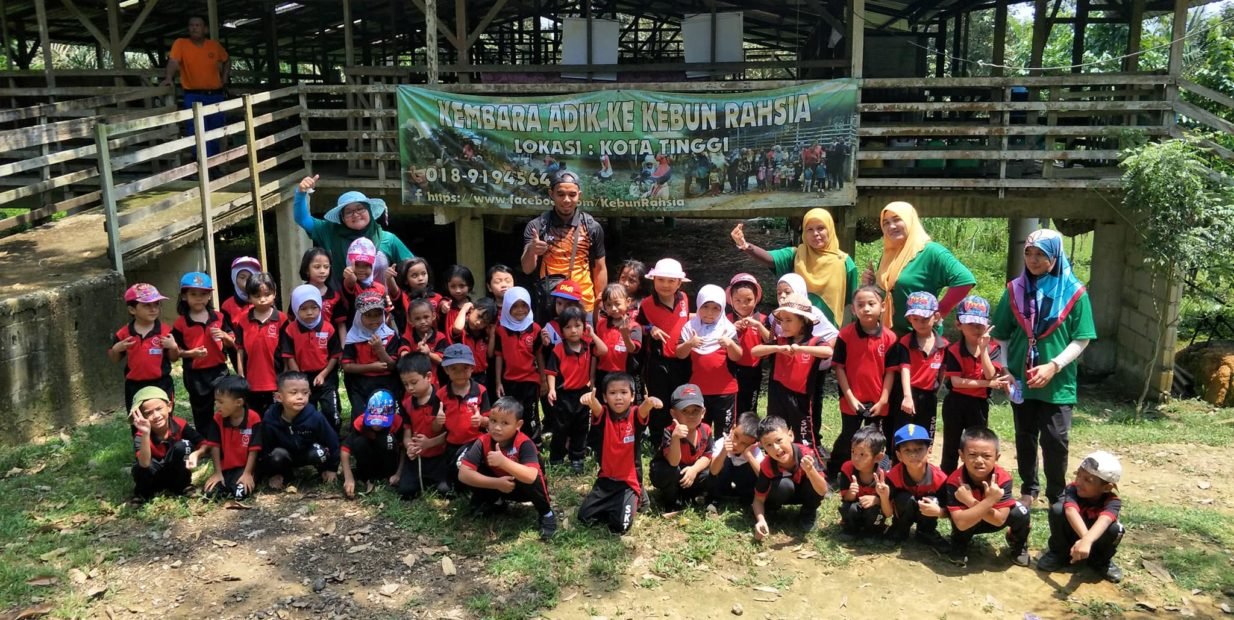 Group of Malaysian kindergarten children and teachers posing in front of a Kebun Rahsia farm banner during an outdoor educational visit in Kota Tinggi, Johor.
