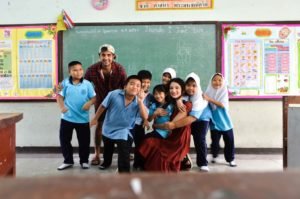 Two international volunteers posing and laughing with a group of Thai primary school children in front of a chalkboard in a classroom in Thailand Pocket money English teaching placements in Thailand with an NGO