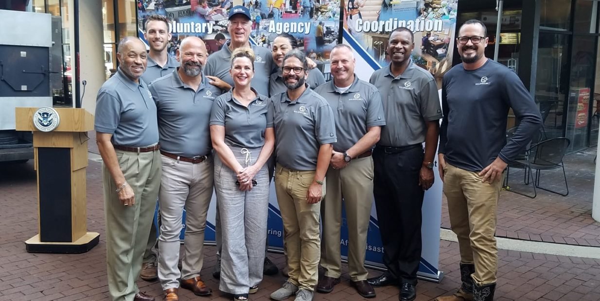 Group of EQUIProject disaster relief volunteers in matching grey shirts posing outside in front of Voluntary Agency Coordination banners.