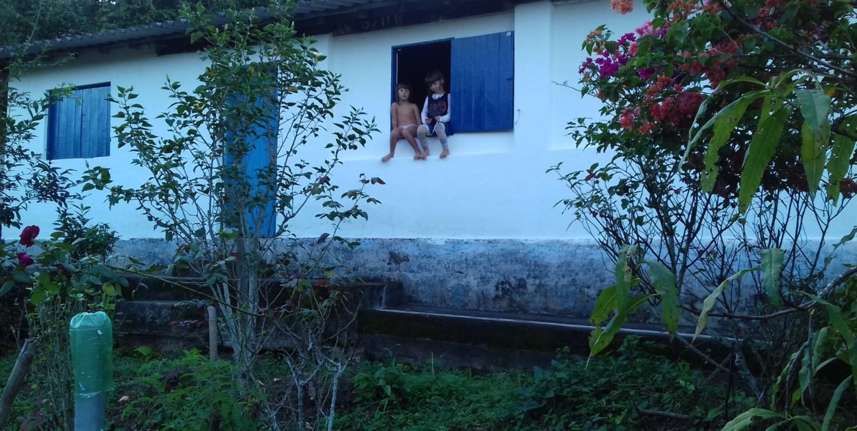 Simple white farmhouse with blue shutters surrounded by garden plants, with two small children sitting in the window, part of an organic farm volunteer project in Brazil.