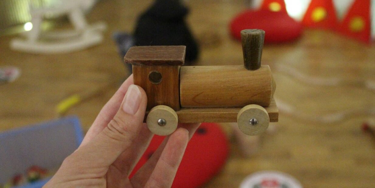 Hand holding a small wooden toy train in a playroom at Little Prince Academy, Göreme (Cappadocia); red cushions and a play tent blurred in the background.