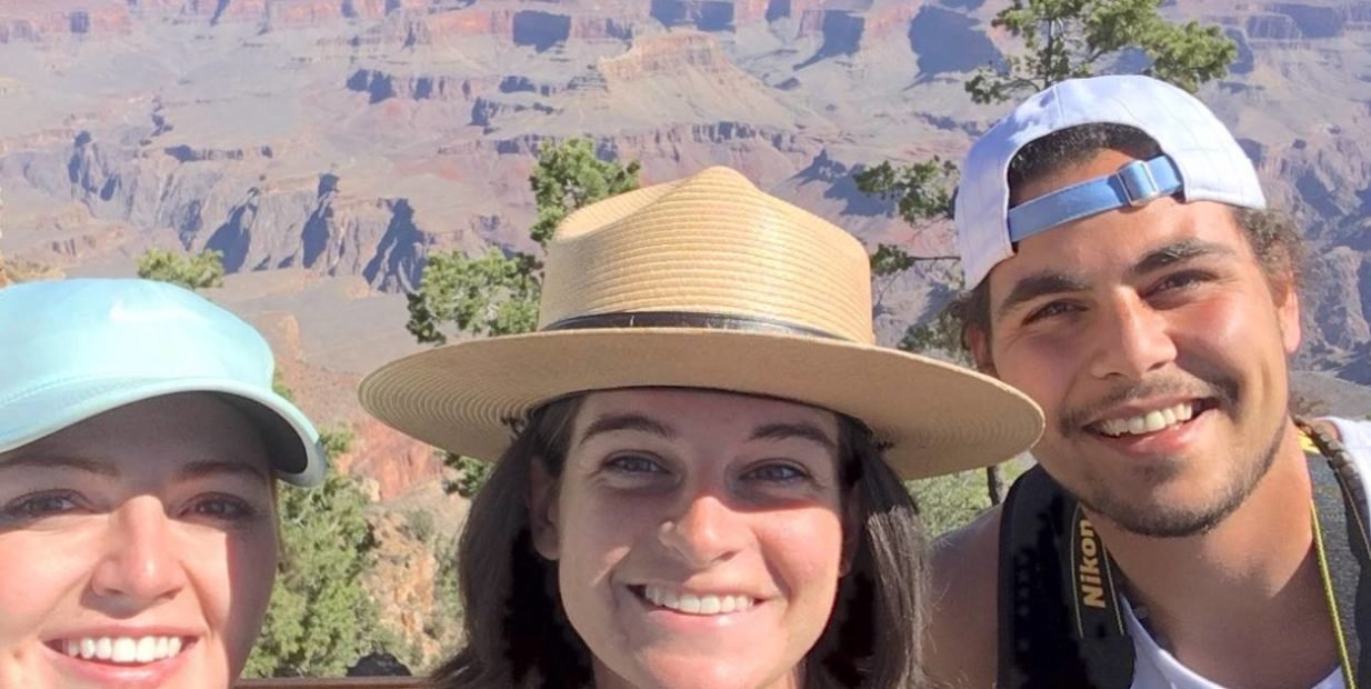 Three smiling young adults taking a selfie at a scenic canyon viewpoint, with colourful rock cliffs, trees and blue sky in the background, representing happy volunteers exploring a US national park.