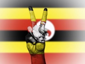 Uganda volunteer projects concept, hand showing a peace sign painted with the Uganda flag colors in front of a blurred Uganda flag background.