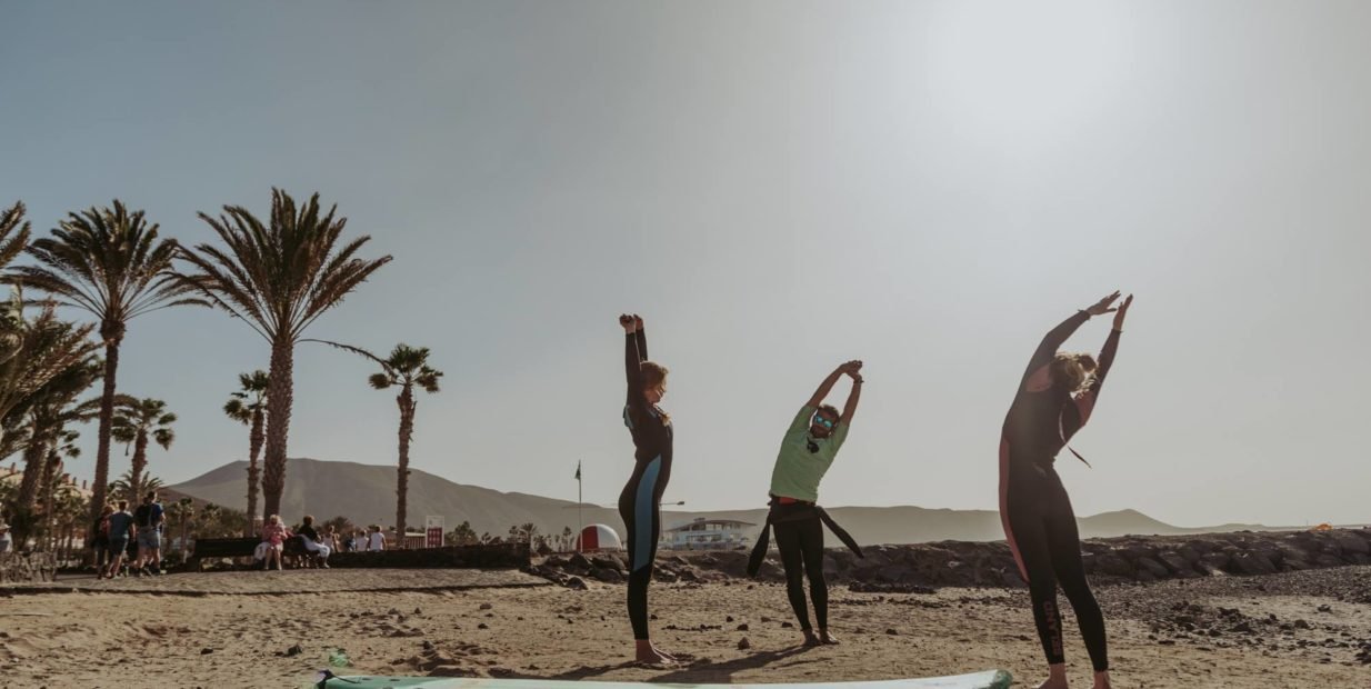 Three surfers in wetsuits stretching beside their surfboards on a sandy beach in Tenerife, with palm trees, mountains and bright sun in the background.