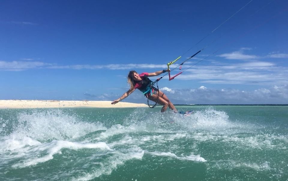 Woman kitesurfing on turquoise water near a sandy island in the Philippines, leaning back as she rides a wave under a clear blue sky.