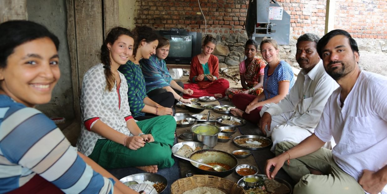 Group of Indian hosts and international volunteers sitting on the floor around metal plates, sharing a simple home cooked meal in a rural village in Maharashtra, India.