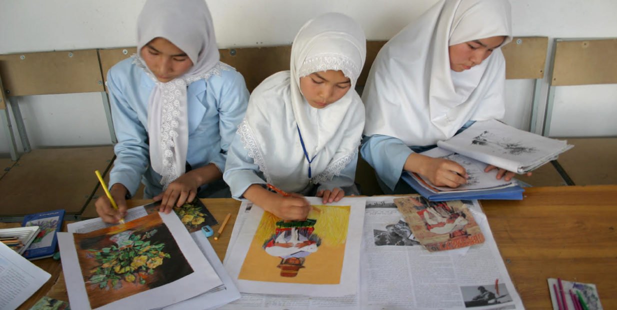 Three Afghan schoolgirls wearing white headscarves sitting at a desk, drawing and studying colorful pictures during an art or English class.