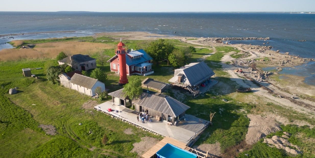 Aerial view of Viirelaid private island in Estonia with a red lighthouse, wooden guesthouses, a small pool and open shoreline surrounded by the Baltic Sea.