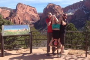 Three friends at a scenic viewpoint near Las Vegas, two holding a third person upside down in a playful pose with red desert cliffs and canyons in the background, showing the fun communal living volunteer experience.