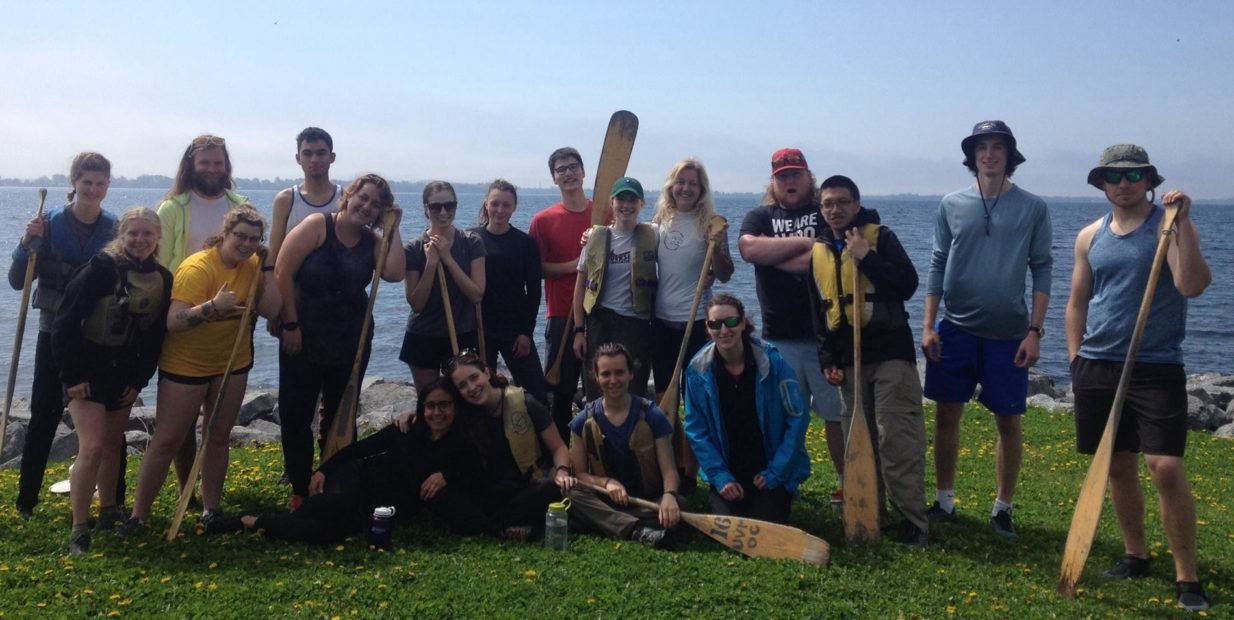 Group of Camp Outlook volunteers standing on green grass by a Canadian lake, holding canoe paddles and smiling at the camera.