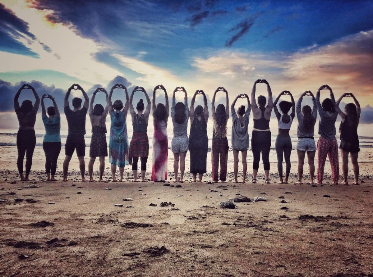 Group of people standing in a line on a Costa Rican beach at sunrise or sunset, facing the sea with their arms raised overhead in a yoga-style pose.