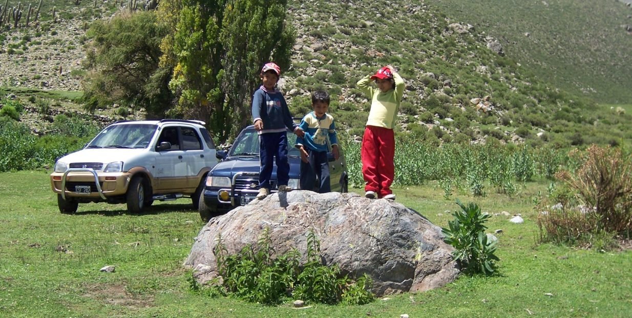 Children standing on a large rock in a green mountain valley in Argentina, with two cars parked behind them and hills in the background near a mountain refuge.