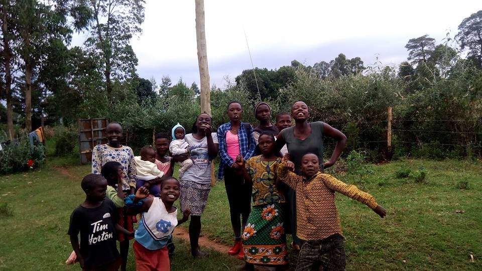 Group of smiling children and women standing together on green grass in rural Kenya, posing for the camera on a cloudy day.