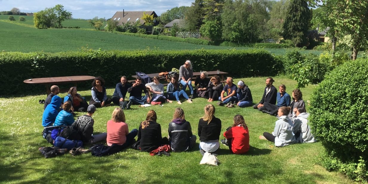 Group of volunteers sitting in a big circle on the grass at a rural retreat centre in Denmark, talking together on a sunny day at Møn Retreat Center