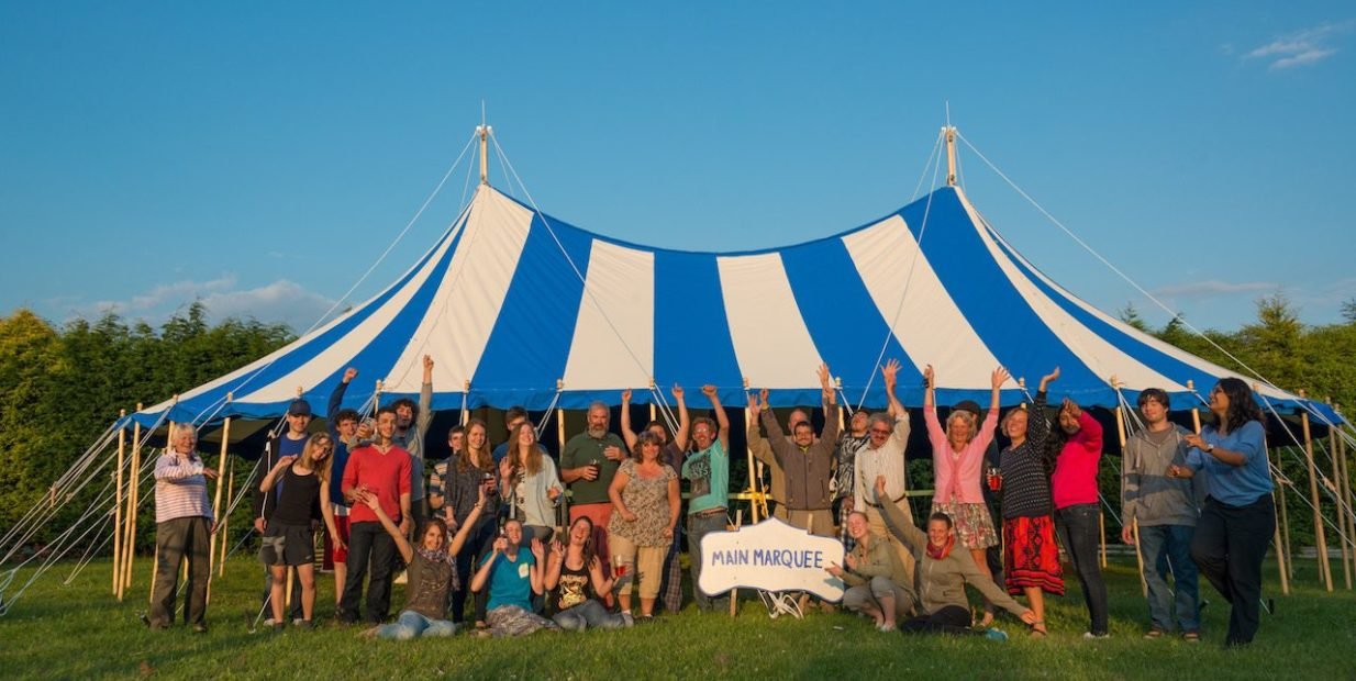 Large group of volunteers cheering in front of a big blue and white striped marquee tent on a sunny green field at Green & Away in England.