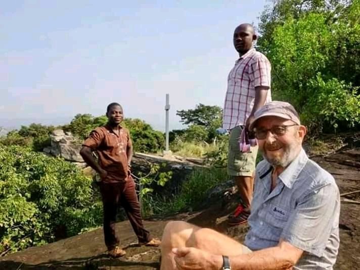 Three men relaxing on a rocky viewpoint surrounded by green hills and trees in rural Ghana on a sunny day.