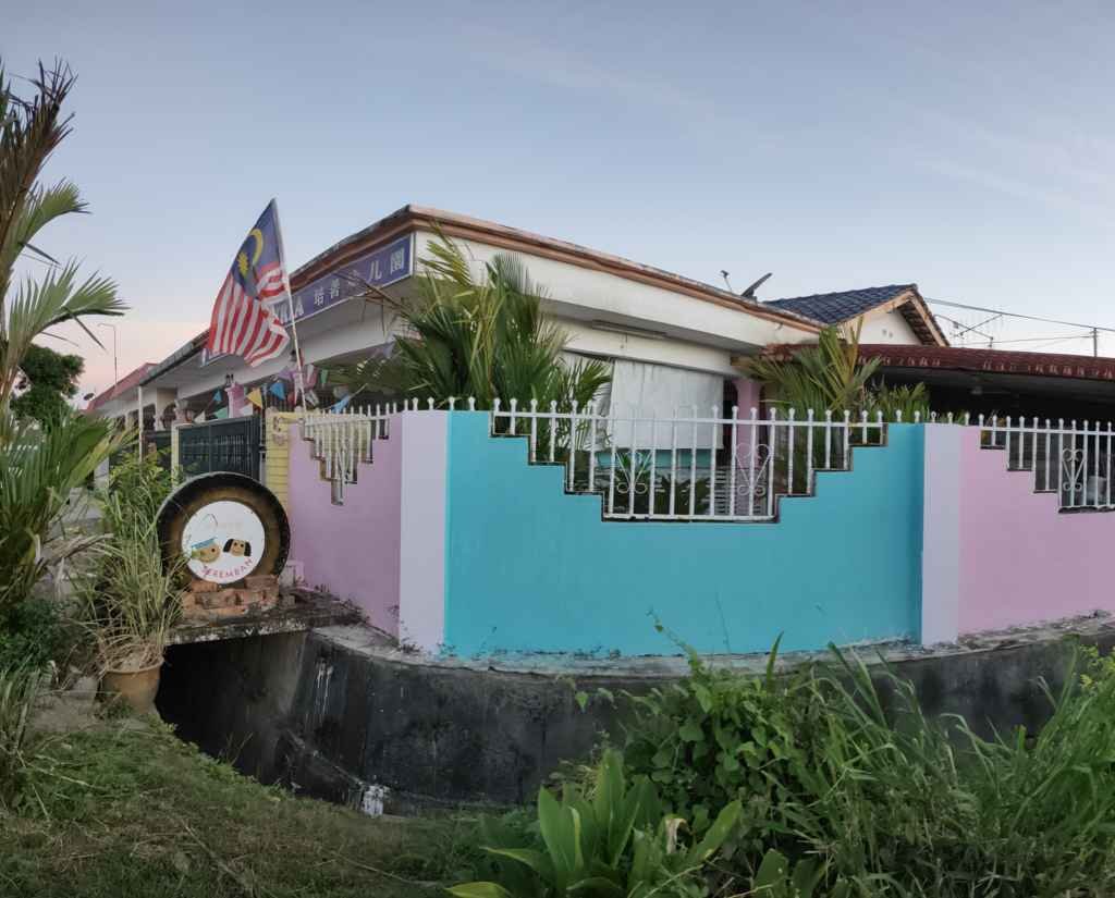 Single-storey kindergarten building in Seremban, Malaysia, with colourful pastel walls, palm plants and a Malaysian flag at the gate.