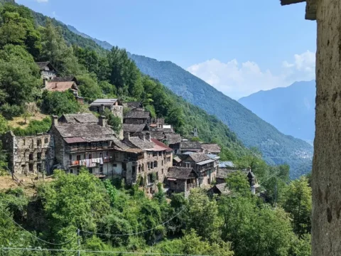 View of Zonca, a small stone village on a green mountainside in northern Italy, surrounded by forest and alpine valleys.