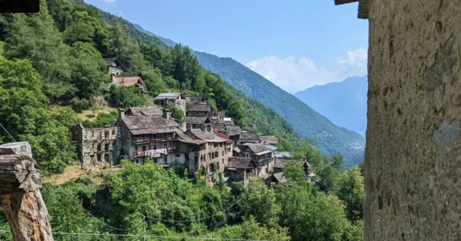 View of Zonca, a small stone village on a green mountainside in northern Italy, surrounded by forest and alpine valleys.