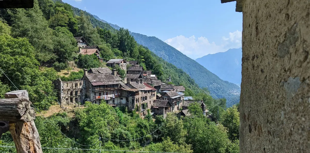 View of Zonca, a small stone village on a green mountainside in northern Italy, surrounded by forest and alpine valleys.