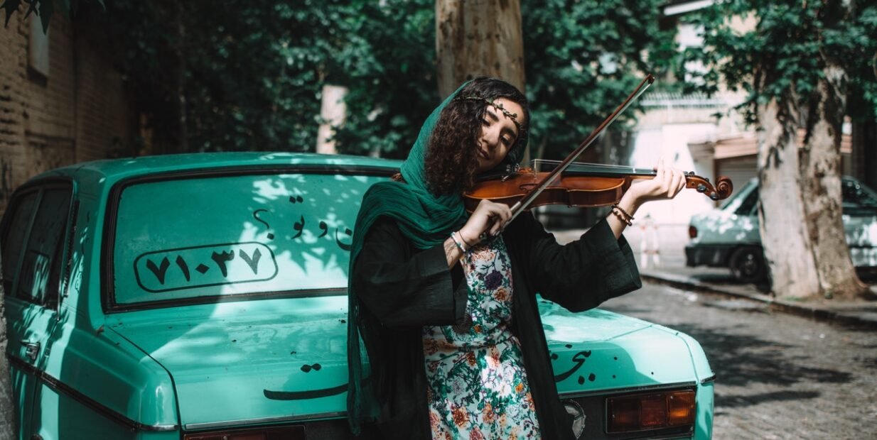 Woman playing a violin on a quiet street beside a green car, a creative, cultural exchange mood for an international student festival opportunity in Iran. Youth programme, volunteering opportunity, cross-cultural projects, Voluntouring.