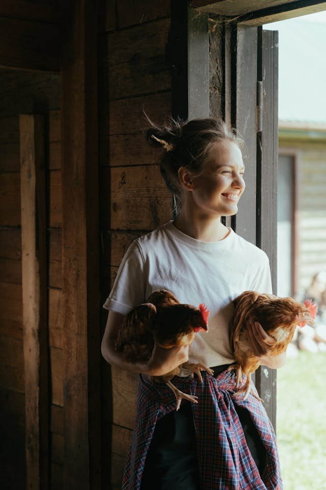 Smiling young woman holding two chickens at the entrance of a wooden farm building, representing volunteer work with animals in a rural setting.