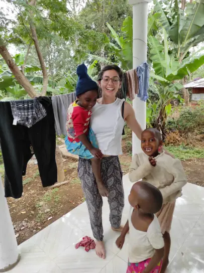 A smiling female volunteer stands on a porch with three children at Tabasamu Orphanage in rural Tanzania. One child is in her arms while others smile beside her, surrounded by banana trees and hanging laundry.