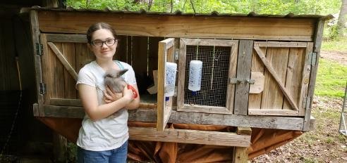 Young farm volunteer holding a rabbit in front of a rustic wooden hutch at Abundant Grace Ranch