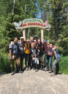 Group of volunteers standing together at the Warren Woessner Bog Boardwalk during a bear sanctuary volunteer project in Minnesota. Outdoor nature setting with forest background. Participants of a wildlife conservation and hospitality-exchange program that offers free food and accommodation while helping protect American black bears