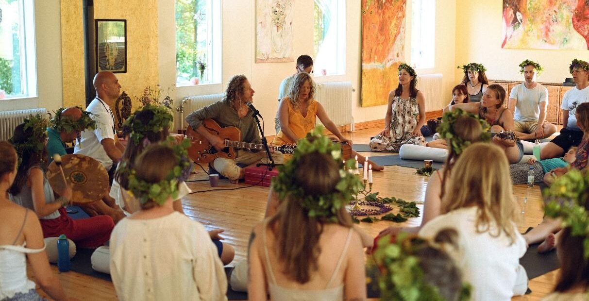 Participants at a Shambala Gatherings spiritual retreat in Sweden sit in a circle wearing flower crowns, engaging in a music and meditation session inside a light-filled hall surrounded by art and nature.