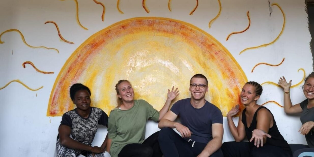 Group of international and local volunteers at A Joy Life, smiling in front of a painted sun mural at A Joy Life project in Tanzania, highlighting community spirit and creative engagement.
