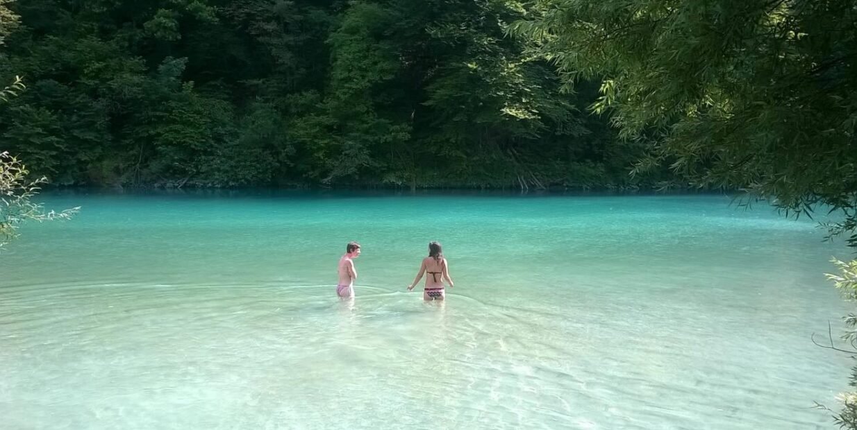Two people wading in a clear turquoise river near Tolmin in Slovenia’s Soča Valley, a calm summer swim spot often linked with the Sajeta festival setting.