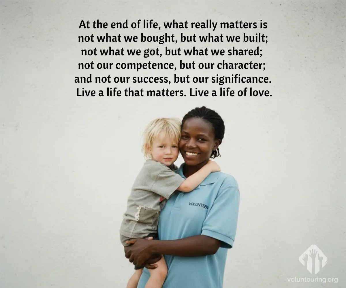 Smiling Black female volunteer in a blue “VOLUNTEER” shirt holding a small blond child against a plain wall. Quote reads, “At the end of life, what really matters is not what we bought, but what we built; not what we got, but what we shared; not our competence, but our character; and not our success, but our significance. Live a life that matters. Live a life of love.”