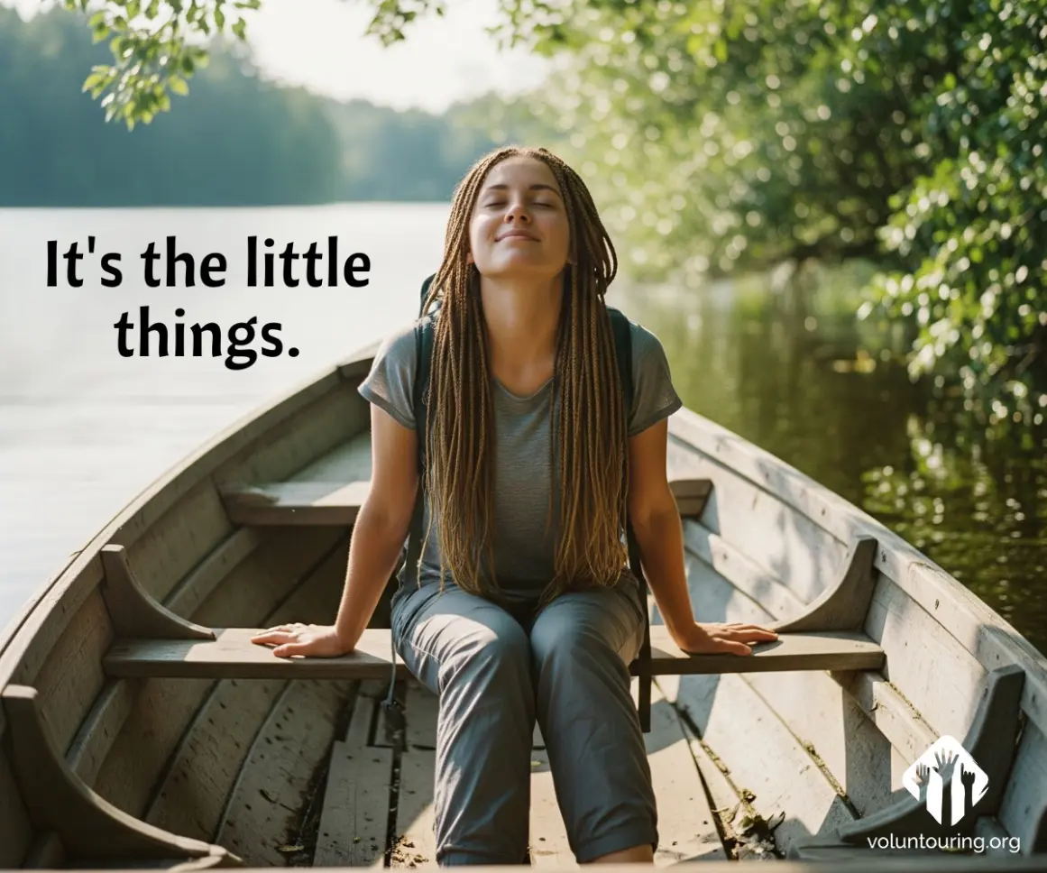 Young woman with long braids relaxing peacefully in a wooden rowboat on a calm river surrounded by trees, with the quote 'It's the little things' and the Voluntouring.org logo.