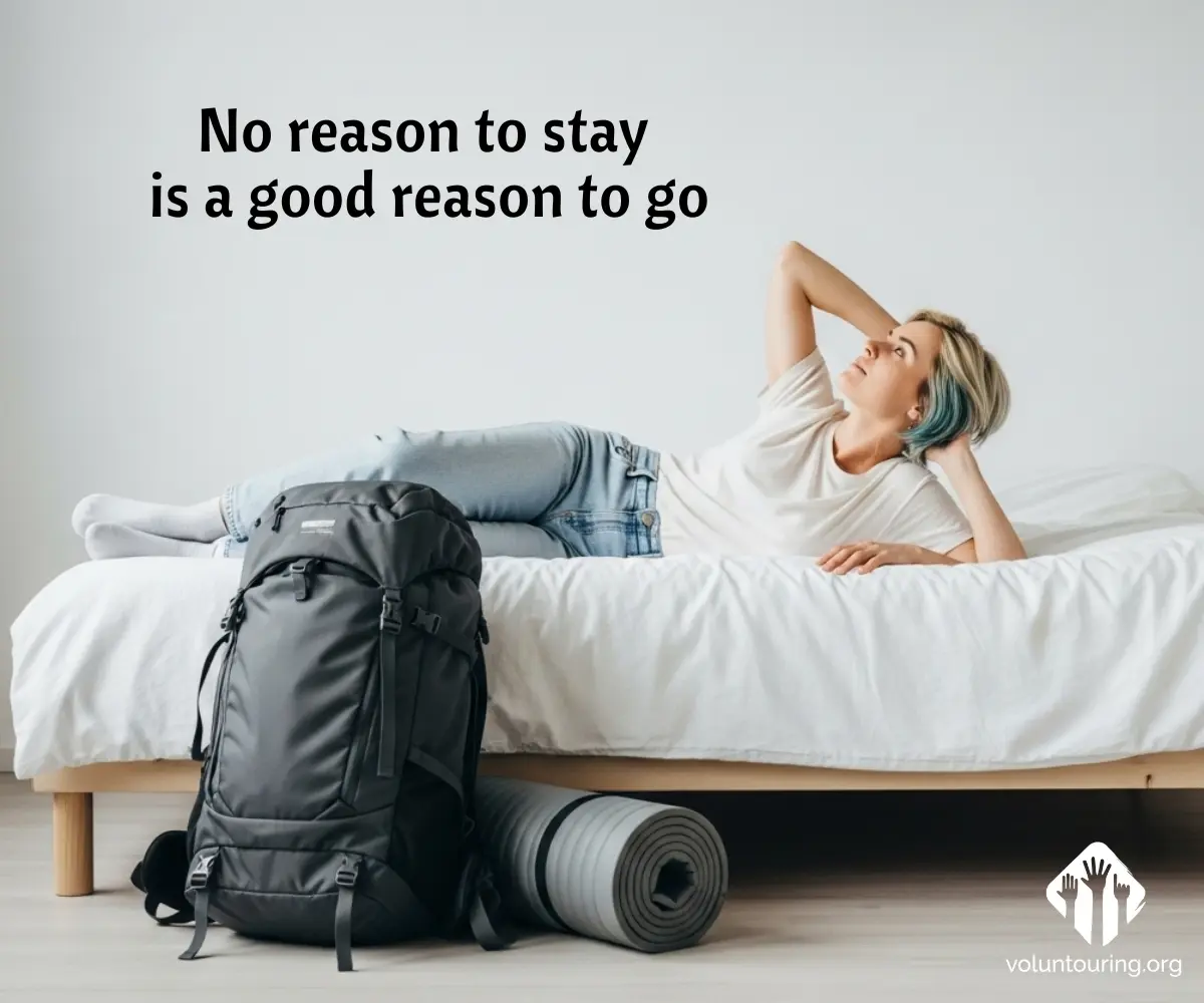 A young woman with short blonde and teal hair lounges on a bed, gazing thoughtfully upward. On the floor next to her are a large black backpack and a rolled-up sleeping mat. The quote “No reason to stay is a good reason to go” appears above her. The voluntouring.org logo is in the bottom-right corner.